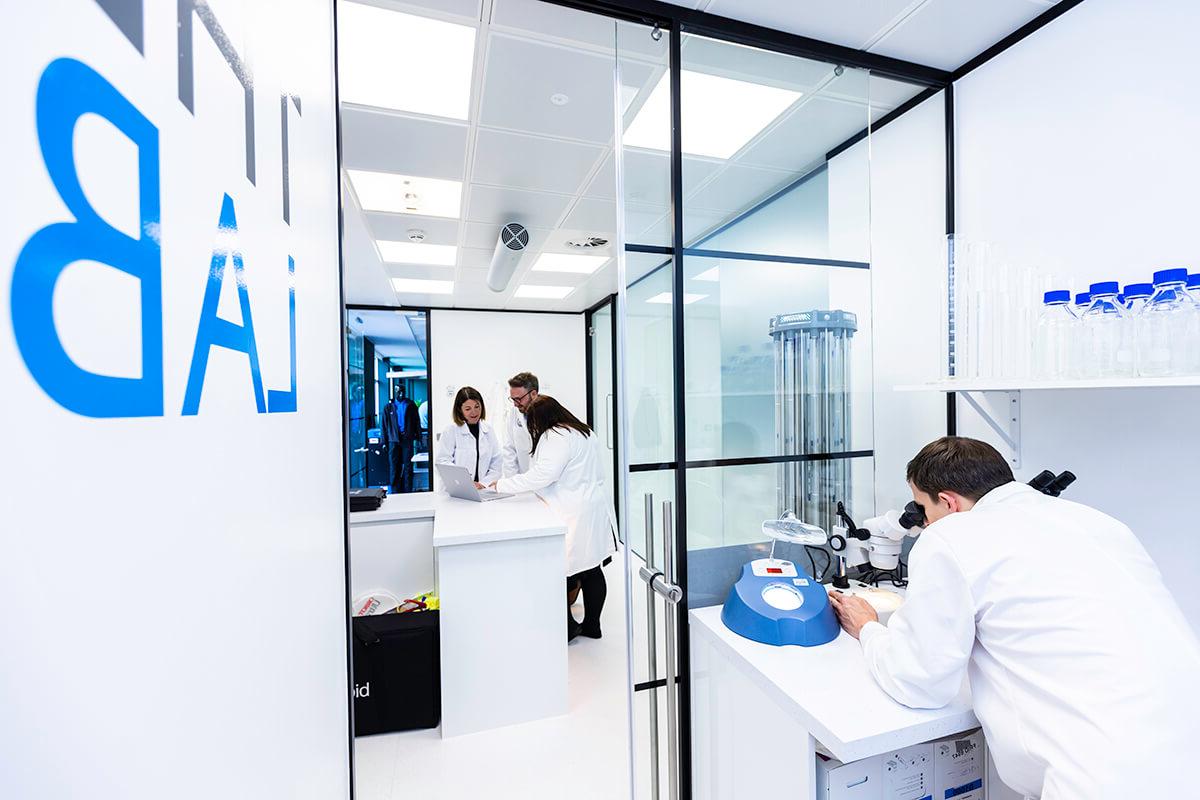 A group of people wearing white lab coats, one looking through a microscope, in a white-walled set of rooms called 实验室 in the Mitie清洁 and Hygiene Centre of Excellence